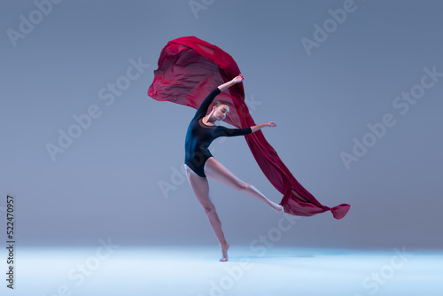 Portrait of young ballerina dancing with deep red fabric isolated over blue grey studio background. Tender wave