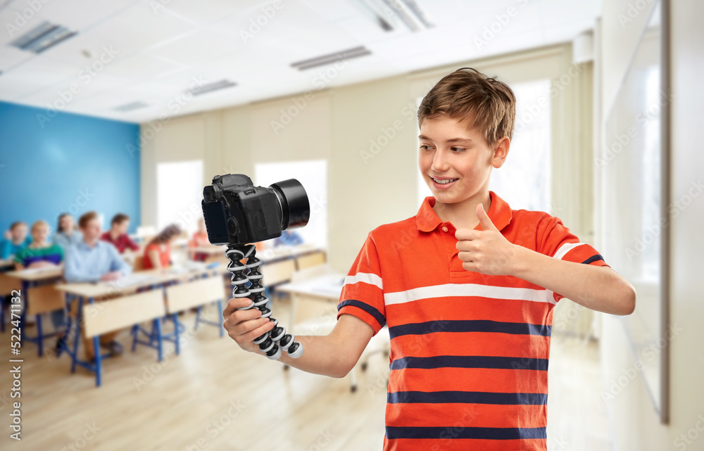 education, school and people concept - happy smiling student boy video blogger with camera and tripod videoblogging and showing thumbs up over classroom background