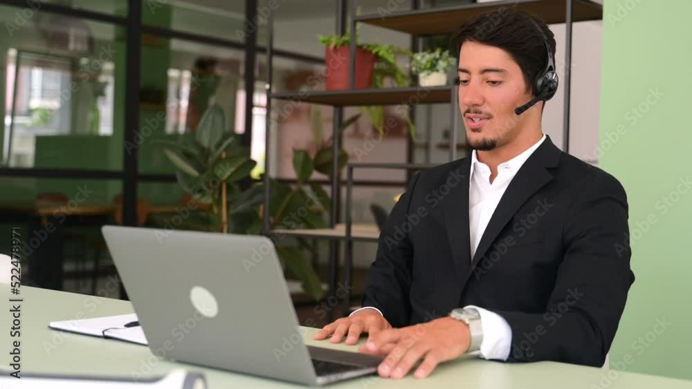 Friendly latin male office employee with a headset using laptop for communication with coworkers or clients, takesa part in video conference. A support officer is talking with a customers, helpline Friendly latin male office employee with a headset using laptop for communication with coworkers or clients, takesa part in video conference. A support officer is talking with a customers, helpline