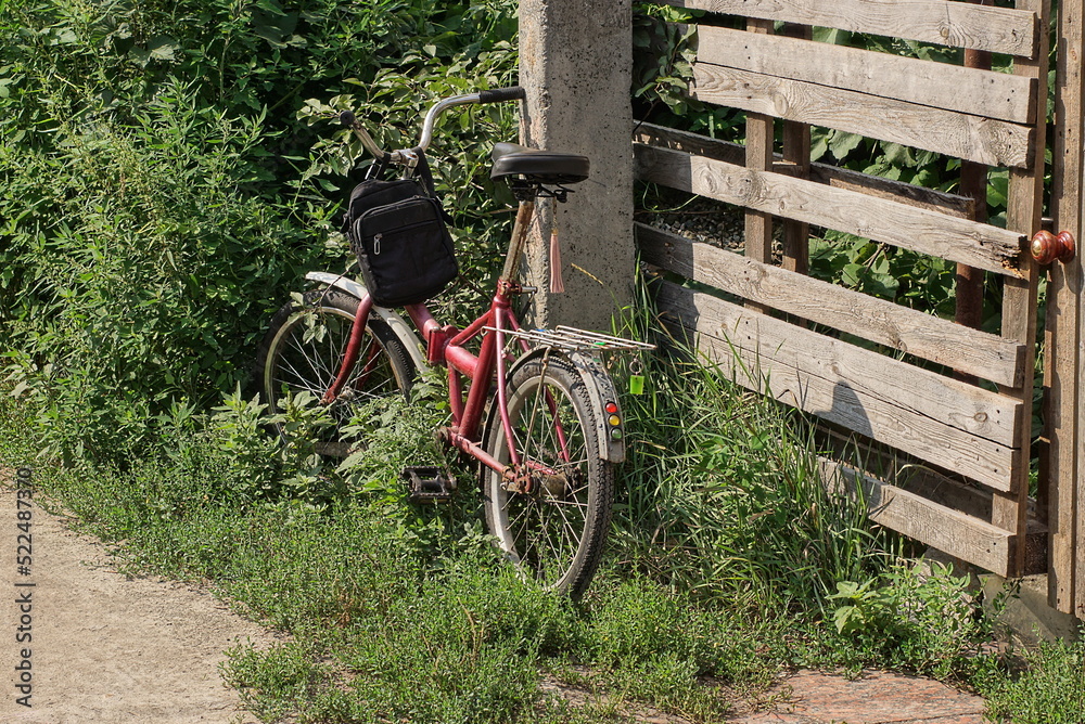 one old red bicycle stands on the street in green grass and vegetation near a gray wooden fence