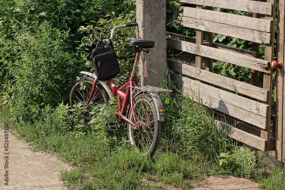 one old red bicycle stands on the street in green grass and vegetation near a gray wooden fence one old red bicycle stands on the street in green grass and vegetation near a gray wooden fence