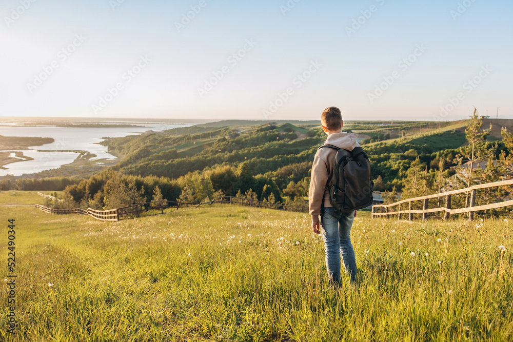 Tourist with backpack standing on top of hill in grass field and enjoying beautiful landscape view. Rear view of teenage boy hiker resting in nature. Active lifestyle. Concept of local travel