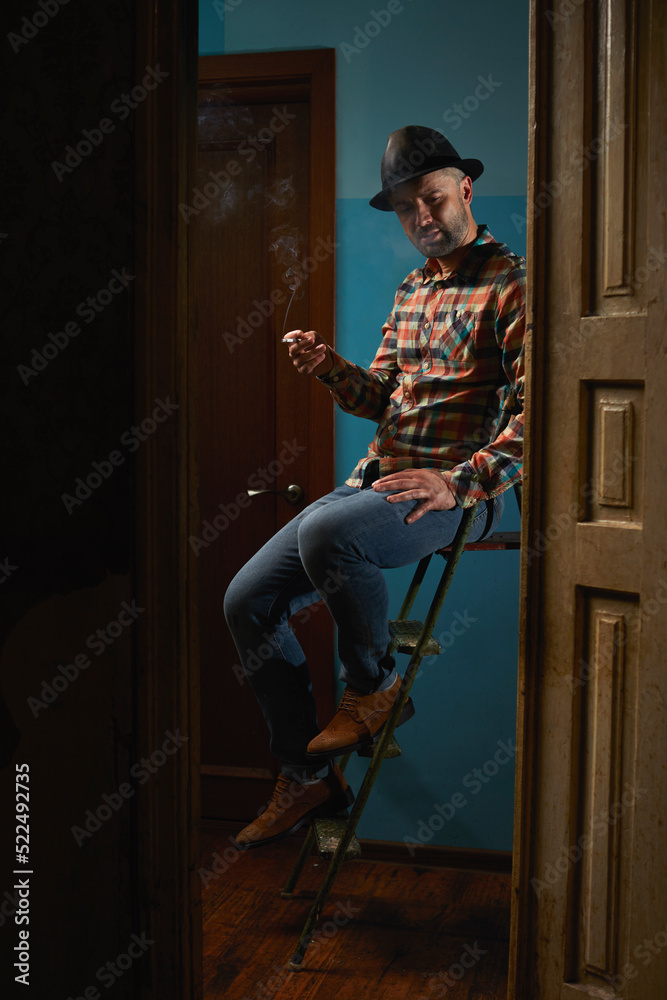 Handsome caucasian man in leather hat siting on ladder for repair and smoking cigarette.