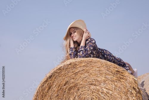 A blonde girl with long hair in a white hat is resting and posing near the sheaves of hay in a field on a sunny day