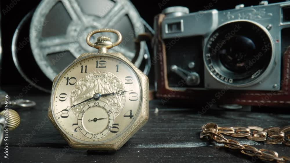 Antique gold and silver pocket watch, camera, gold chain and vintage film reel on black background. Retro pocket watch with ticking second hand. Vintage objects are laid out on dark table. Close up. Antique gold and silver pocket watch, camera, gold chain and vintage film reel on black background. Retro pocket watch with ticking second hand. Vintage objects are laid out on dark table. Close up.