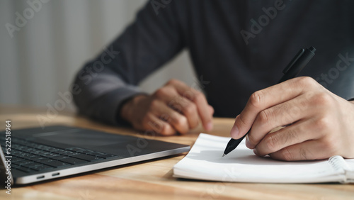 left handed man writes in a notebook on the table with laptop computer