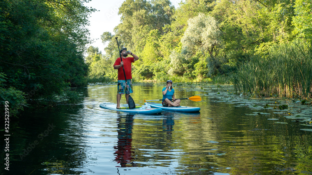 A man with a beard and a woman stand on paddle boards on the river and drink. Summer time