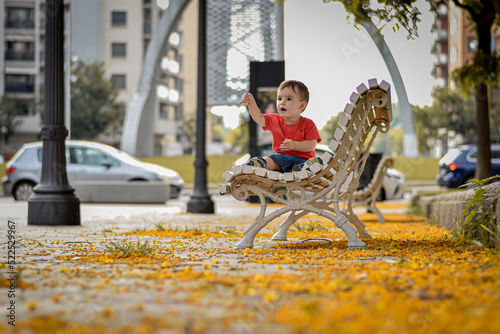 niño sorprendido sentado en un banco de madera en medio de la rambla de una ciudad llena de flores caidas de los árboles y con una rama en la mano  photo