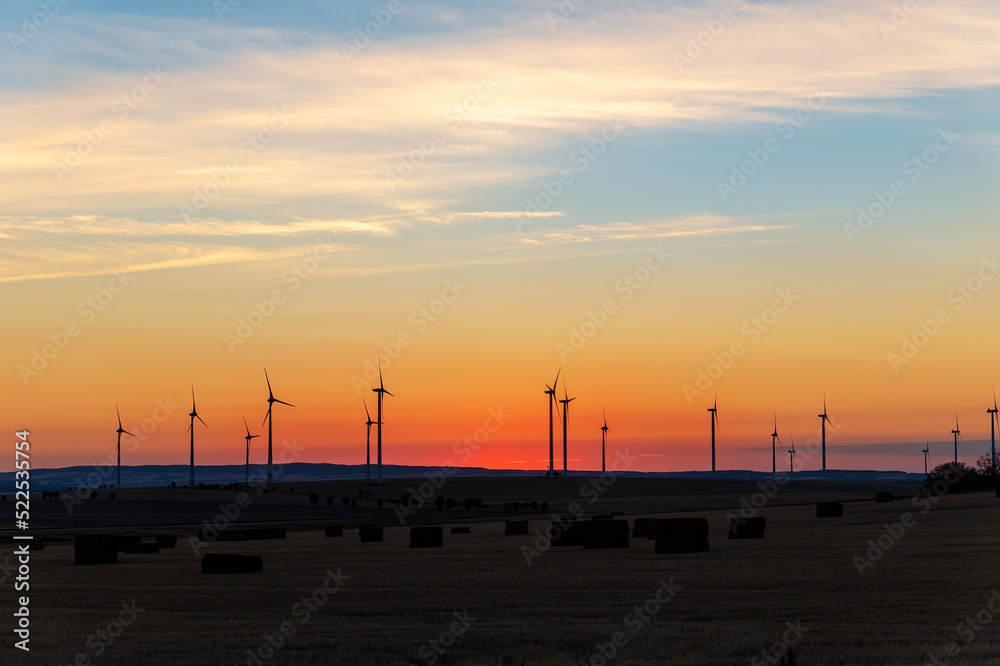 Naklejka premium Panoramic scenic landscape view new modern wind turbine farm power generation station against fiery warm sunset sky field. Clean sustainable zero emission alternative electricity windfarm industry