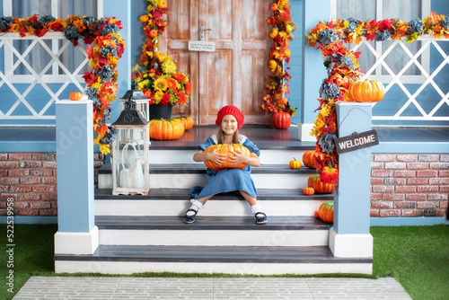 Little happy girl holding pumpkin and smiling at camera while sitting on house porch and waiting to trick or treat on Halloween. Child is sitting next to pumpkins near door of house. Autumn home decor © stock_studio
