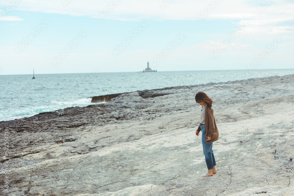 woman walking by rocky sea beach in wet jeans lighthouse on background