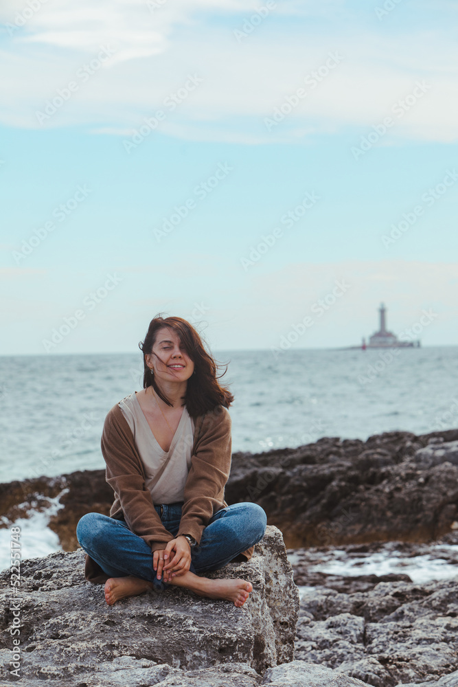 woman sitting by rocky sea beach in wet jeans lighthouse on background