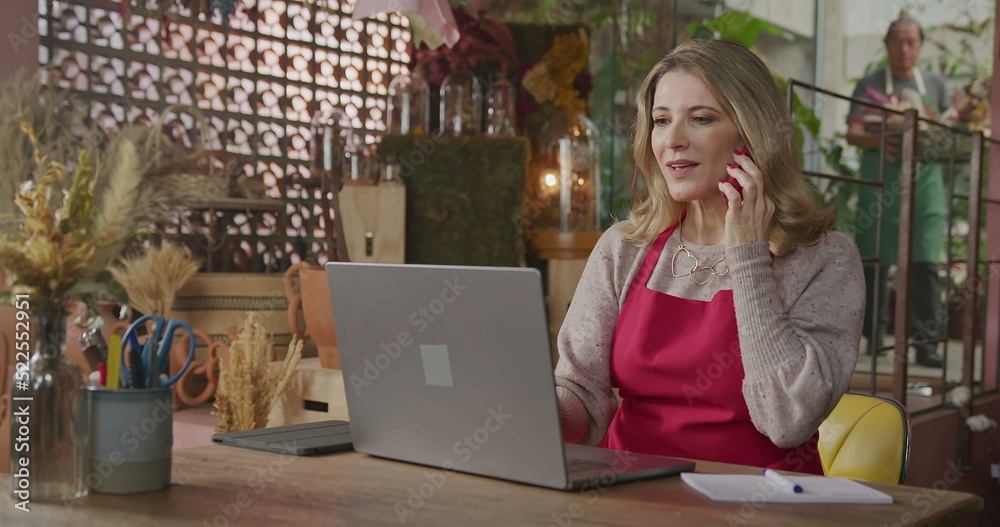 Owner of small business taking online orders in front of laptop computer. Female entrepreneur of local flower shop talking with customer or partner online wearing apron talking on phone Owner of small business taking online orders in front of laptop computer. Female entrepreneur of local flower shop talking with customer or partner online wearing apron talking on phone