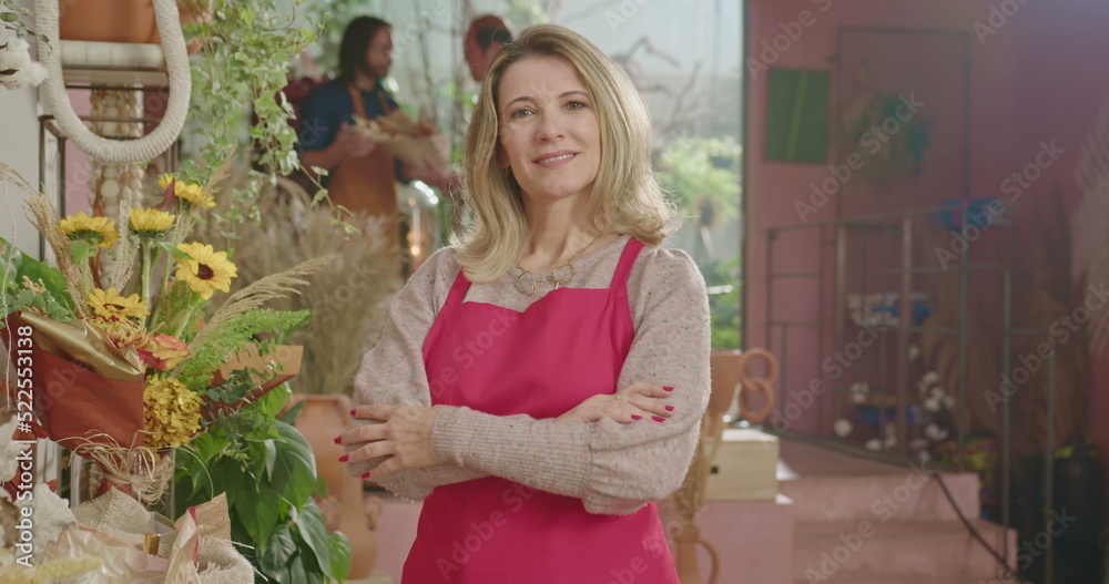 Portrait of a female business store owner smiling at camera with arms crossed with people working in the background. A middle aged businesswoman wearing apron. Small business concept Portrait of a female business store owner smiling at camera with arms crossed with people working in the background. A middle aged businesswoman wearing apron. Small business concept