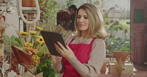 One happy middle aged female entrepreneur holding tablet standing inside flower shop checking for online orders with digital modern device. 
