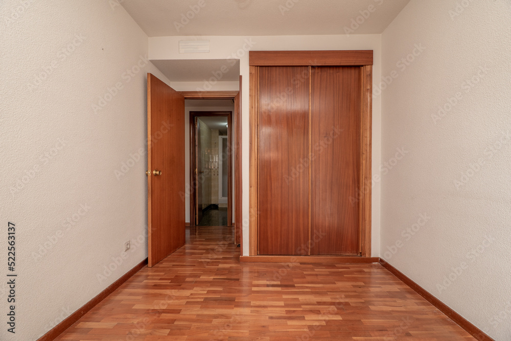 Obraz premium Empty bedroom with bright reddish parquet, matching fitted wardrobes with sliding doors, white gotelet painted walls and cloakroom to the rear