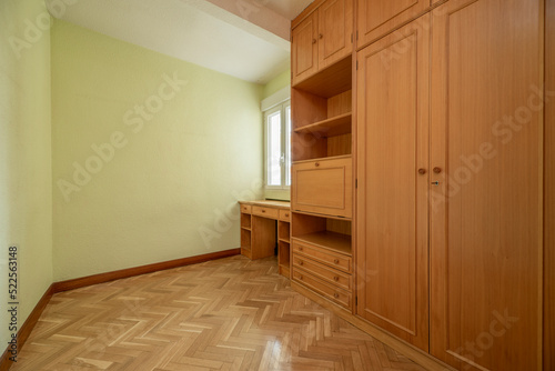 Empty bedroom with herringbone oak parquet, fitted wardrobes, desk and chest of drawers completely covering light wooden wall and light green painted wall