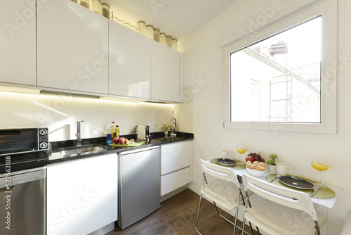 Apartment with open kitchen with white wooden furniture with stainless steel appliances and black marble countertop, breakfast table next to the window.