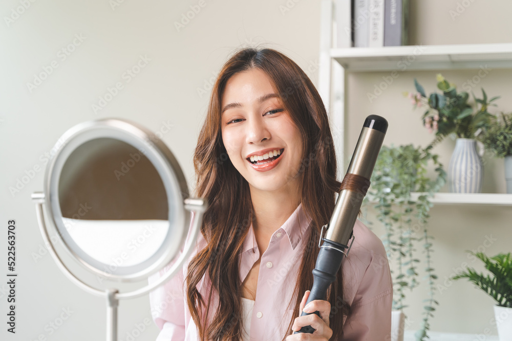 women hairdo makeup routine, Young woman looking at the mirror and using iron hair curling curly long hair at home. - obrazy, fototapety, plakaty 