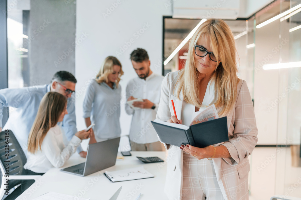 Woman in glasses standing in the front of her colleagues. Group of professional business people is in the office Woman in glasses standing in the front of her colleagues. Group of professional business people is in the office