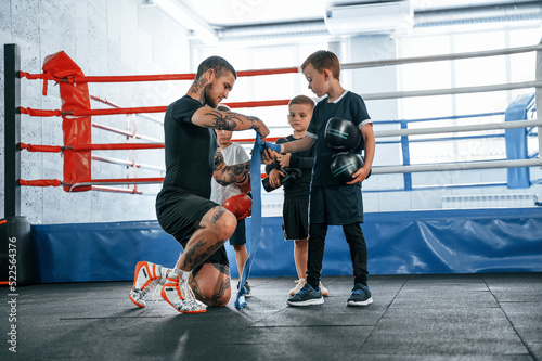 Wearing blue hand wraps. Young tattooed coach teaching the kids boxing techniques