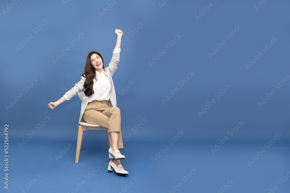 Obraz premium Attractive beautiful Asian woman sitting on white modern chair and hands up raised arms from happiness isolated on blue background, Excited businesswoman winner success concept
