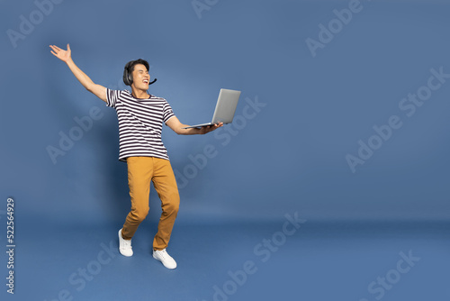 Happy young Asian pro gamer with headphones holding computer laptop and win in online video game isolated over blue background, Full body composition, Cyber sportsman playing tournaments concept