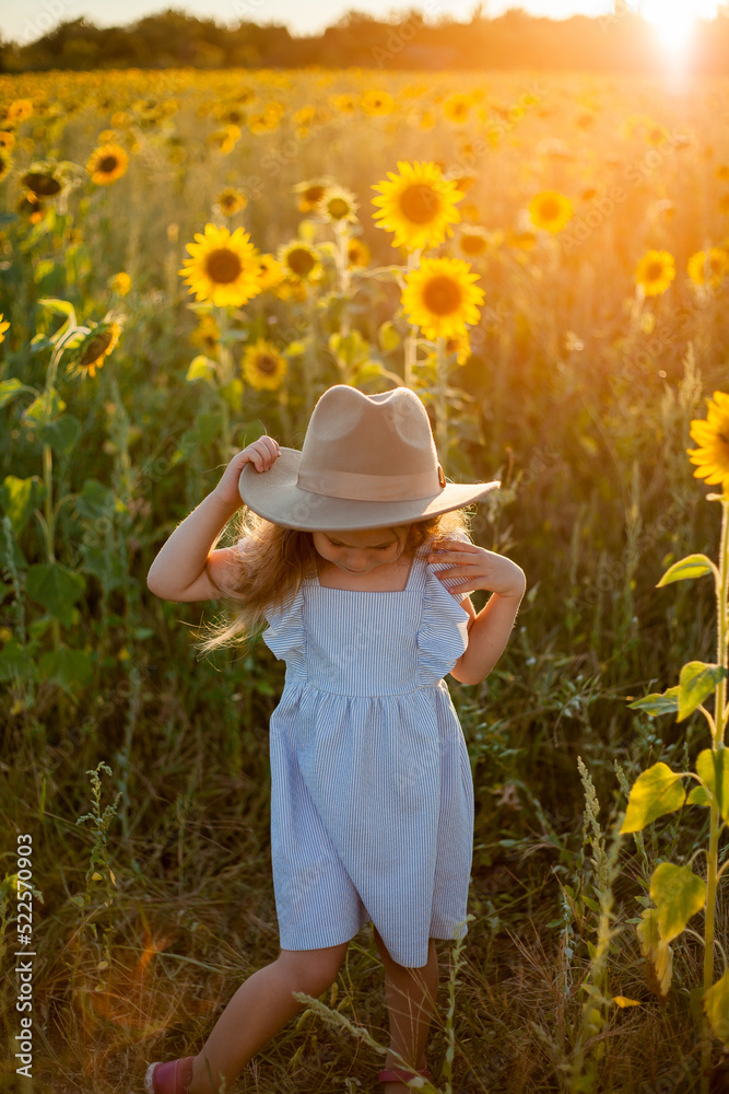 Adorable little girl 4 years old with curly long blond hair in a blue sundress and hat in a sunflower field. Summer portrait of a happy child. Sunset.