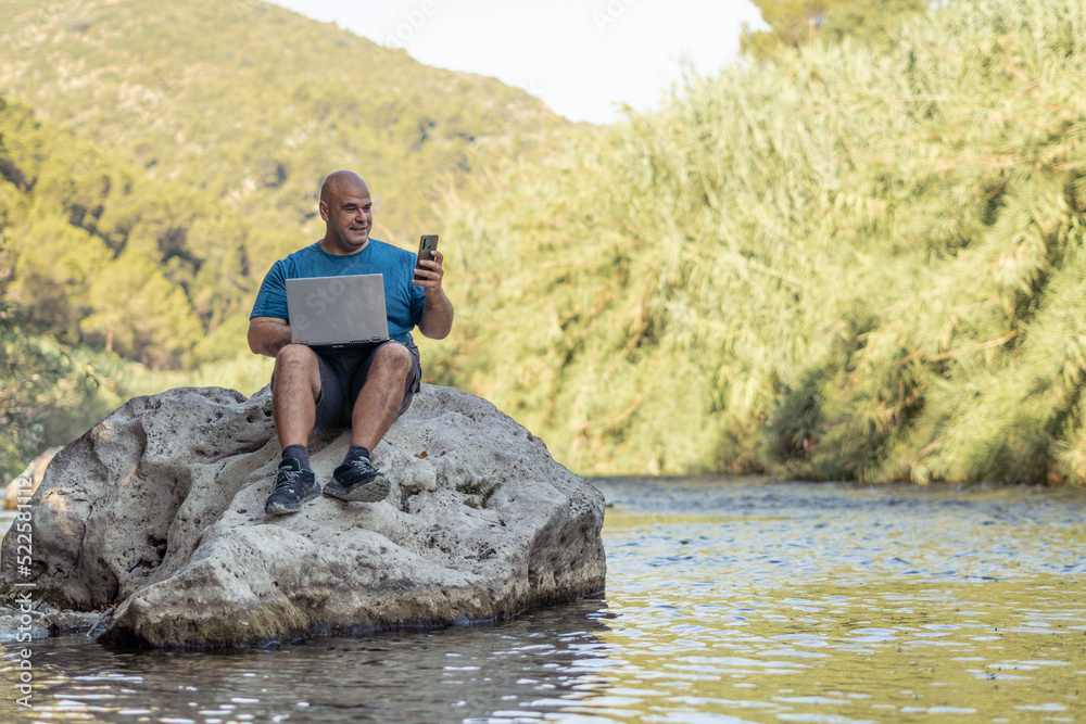 Digital nomad man working with a laptop sitting on a rock above the water of a beautiful and calm river. Digital nomad man working with a laptop sitting on a rock above the water of a beautiful and calm river.