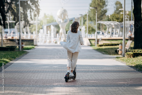 Beautiful young woman in white suit is riding to work on her electric scooter in city parkland © bondvit