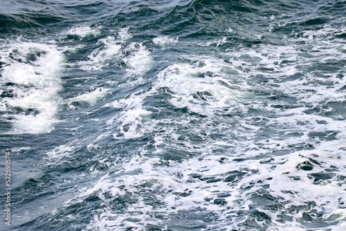 Stern waves with white foam tips on greyish blue sea water, photo taken from aboard ship. Selective focus
