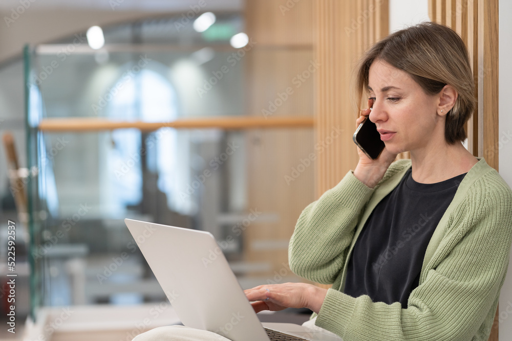 Serious woman holding smartphone discussing project details while working online on laptop in coworking space, talking with client via cellphone, businesswoman with mobile phone looking at pc screen Serious woman holding smartphone discussing project details while working online on laptop in coworking space, talking with client via cellphone, businesswoman with mobile phone looking at pc screen