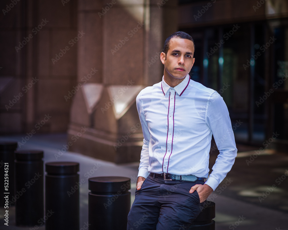Young Man wearing white shirt, two hands putting in pockets, sitting on metal pillar on old street in New York City, relaxing, thinking.