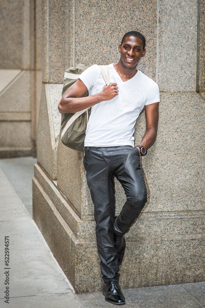 Street Fashion. Young African American man wearing white V neck T shirt, black pants, leather shoes, carrying shoulder bag, standing against column outside in New York City, smiling, looking at you.