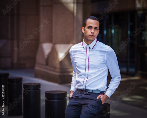 Obraz na płótnie Young Man wearing white shirt, two hands putting in pockets, sitting on metal pillar on old street in New York City, relaxing, thinking. w salonie