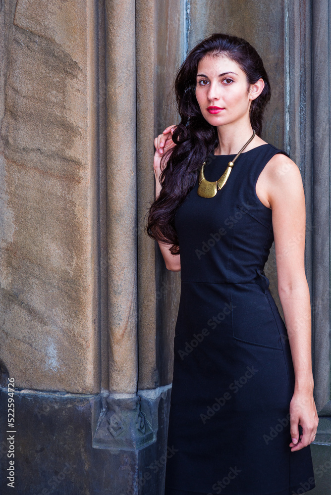Young pretty woman wearing black work dress, necklace with golden pendant, standing outside against old style wall in New York City, looking forward.