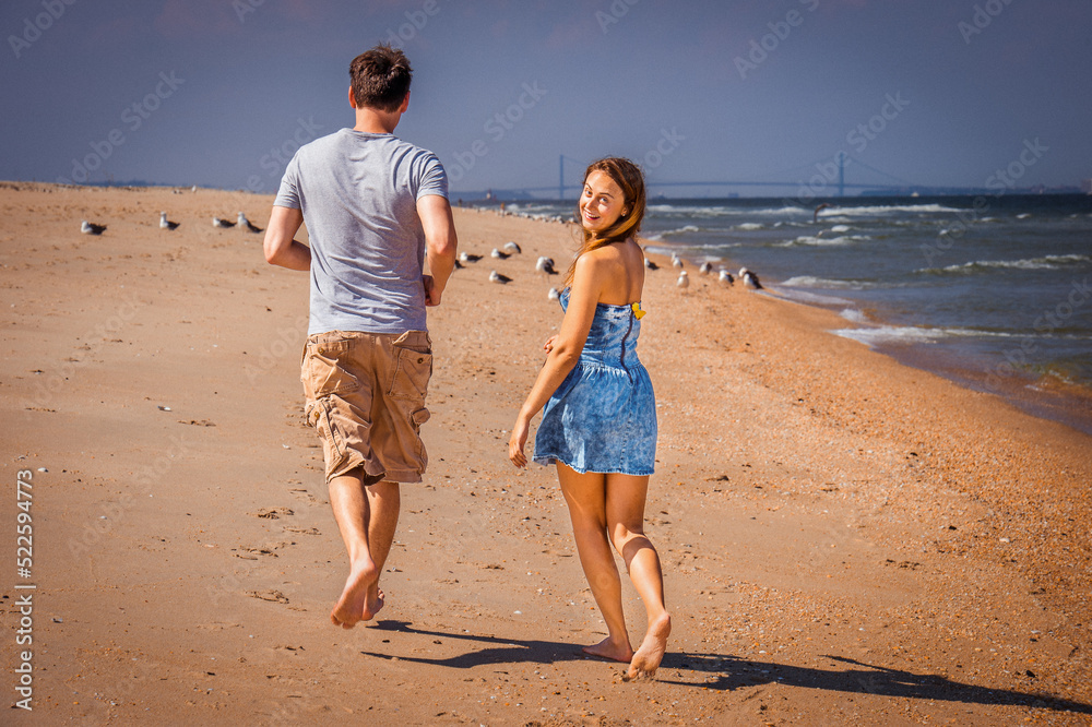 Young couple running on Sandy Hook Beach, New Jersey, USA. Dressing in summer beach outfits, barefoot, girl and guy running, jogging, girl turning back, looking at you..