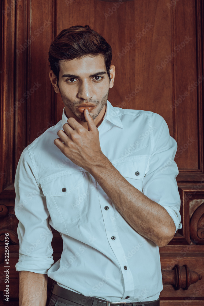Young man wearing white long sleeve shirt, standing by an old style office door, raising arm, thumb touching lip, into deeply thinking..
