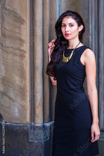 Obraz na płótnie Young pretty woman wearing black work dress, necklace with golden pendant, standing outside against old style wall in New York City, looking forward. w salonie