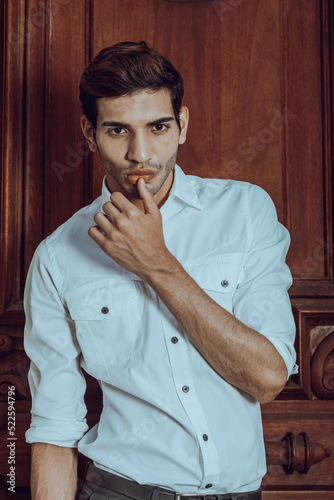 Obraz na płótnie Young man wearing white long sleeve shirt, standing by an old style office door, raising arm, thumb touching lip, into deeply thinking.. w salonie