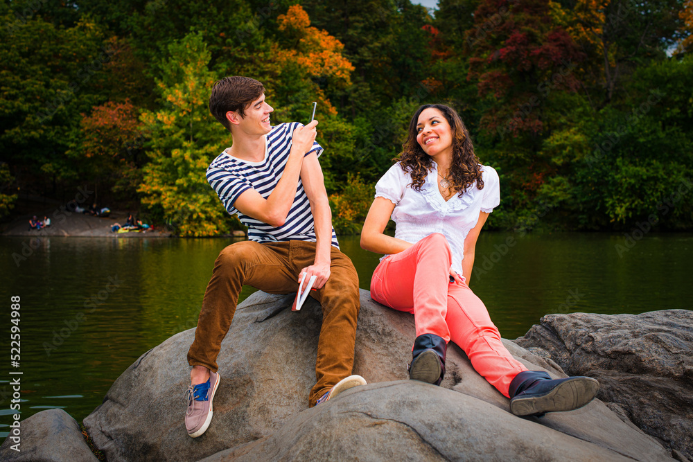 Young couple making fun by a lake in a park. Guy wearing a striped t shirt, brown pants, sneakers, holding a book, taking a picture. Girl dressing in white top, red pants, boot shoes, smiling. .