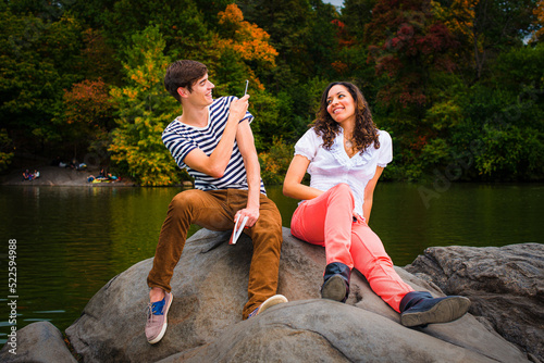 Obraz na płótnie Young couple making fun by a lake in a park. Guy wearing a striped t shirt, brown pants, sneakers, holding a book, taking a picture. Girl dressing in white top, red pants, boot shoes, smiling. . w salonie