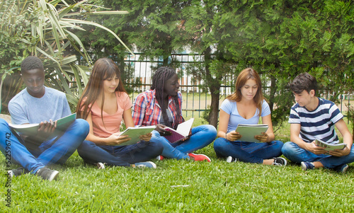 Five teens hang out in a park. Students of multi ethnic classroom seated on the grass doing homework