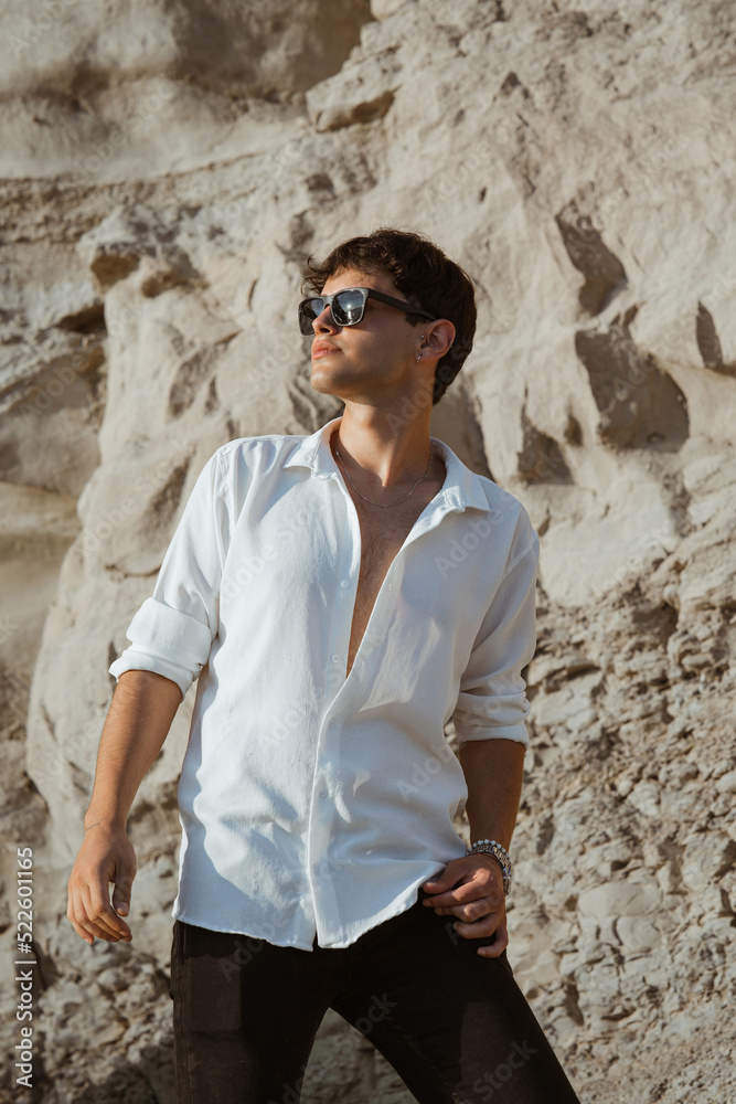 Portrait of handsome sexy young man in white shirt and sunglasses posing staying on sand stones on the beach near sea looking away.