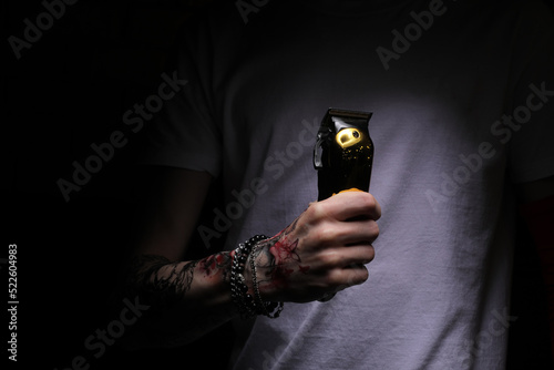 hand of young barber man with stylish tattoo in white t shirt holding electric hair clipper, razor for hairdressers on dark background. Barbershop beauty salon. The barber clippers instead of scissors