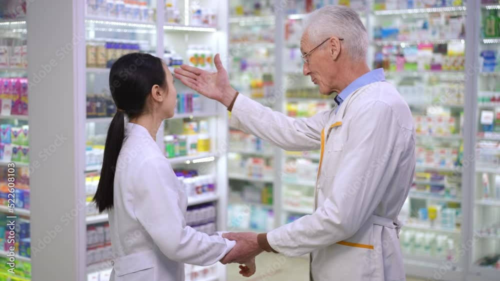 Side view senior Caucasian man and young Asian woman in uniform shaking hands talking in pharmacy leaving. Intelligent professional confident colleagues handshaking in drugstore walking away Side view senior Caucasian man and young Asian woman in uniform shaking hands talking in pharmacy leaving. Intelligent professional confident colleagues handshaking in drugstore walking away