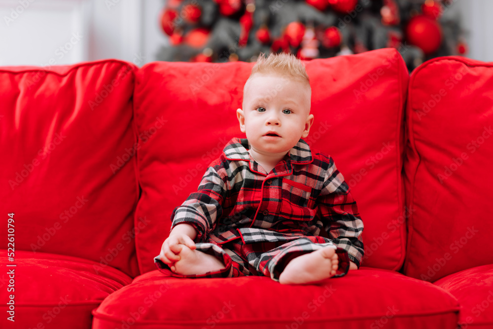 adorable little baby boy in a red christmas outfit sitting on red sofa in front of a christmas tree. Beautiful little baby boy celebrating Christmas.Family on Christmas eve at fireplace. adorable little baby boy in a red christmas outfit sitting on red sofa in front of a christmas tree. Beautiful little baby boy celebrating Christmas.Family on Christmas eve at fireplace.