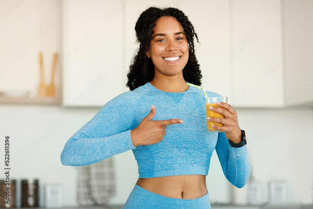 Happy african american lady in sportswear pointing finger at glass of orange juice, standing in kitchen and smiling - obrazy, fototapety, plakaty 