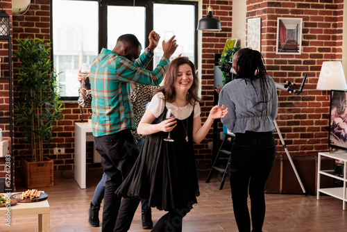 Multiethnic group of closed friends dancing together in living room at wine party while having fun. Young adult people enjoying disco music at home while celebrating friendship anniversary event.
