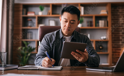 Busy concentrated mature chinese man at table with laptop uses tablet for work in home office interior